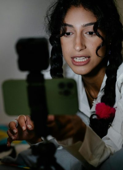 A young woman with braided hair records a video using her smartphone and laptop indoors.